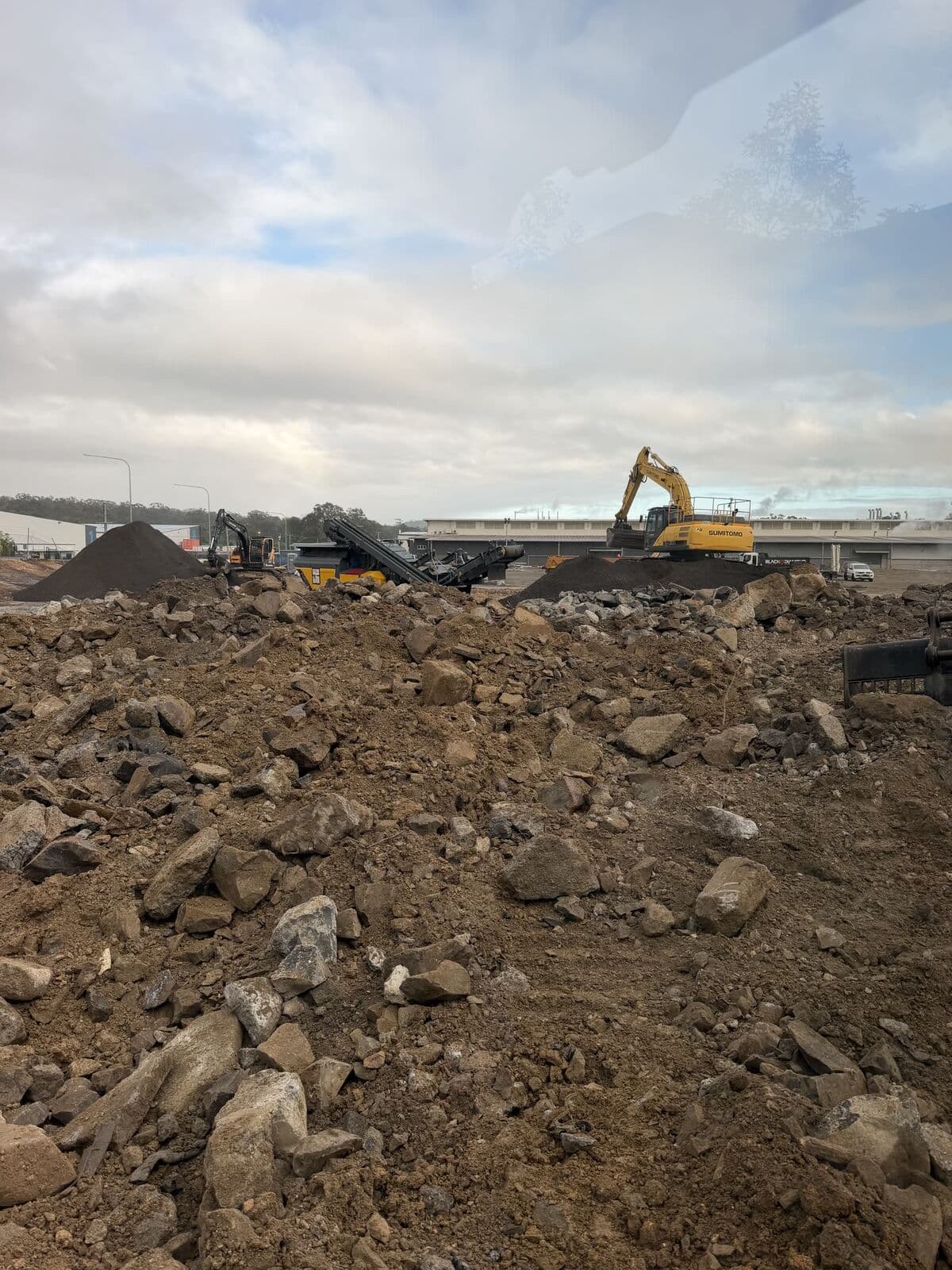 View from an excavator cab across broken concrete and rock, with a Sumitomo excavator and a tracked screening plant working stockpiles on an industrial site